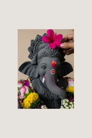 A dark gray Ganesha statue adorned with a bright pink hibiscus flower on its head, surrounded by colorful flowers, with a hand placing the flower on the statue.