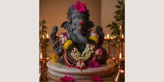 A dark gray Ganesha statue adorned with colorful floral garlands and a pink flower on his head, seated on a marble-like pedestal with lit oil lamps in the background.