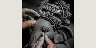 Close-up of a sculptor's hands carefully applying clay or wax to the forehead of an elephant-headed deity statue, with detailed carvings on the crown visible against a dark background.