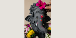 A dark gray Ganesha statue adorned with a bright pink flower on his crown, surrounded by colorful marigold flowers, with a hand placing the flower on his head.